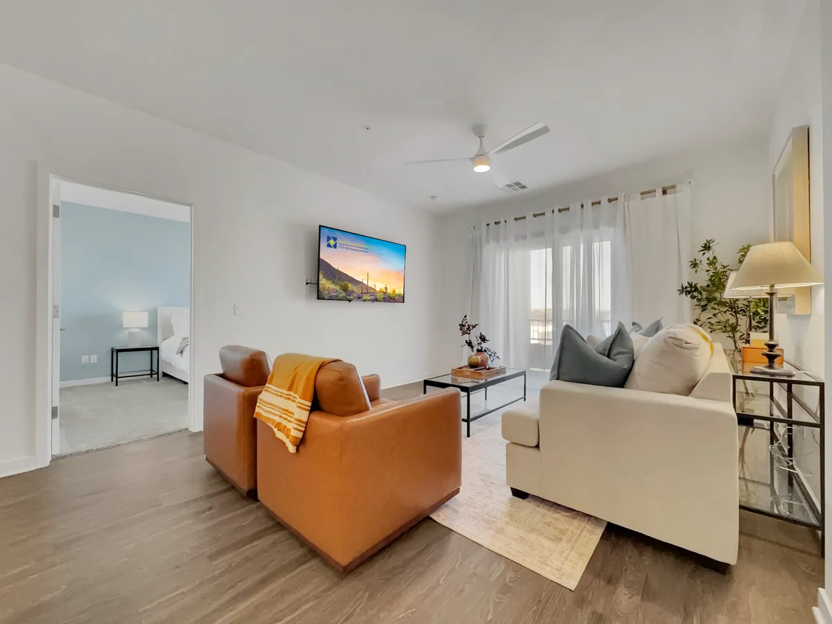 A warm, open-plan living area showcasing a comfortable seating arrangement facing a large television. Generous natural light streams in through expansive windows, while decorative plants and modern touches create a relaxed yet sophisticated ambiance.