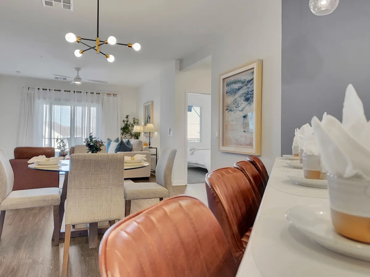 This image features a well-lit dining area with a dining table surrounded by bar stools, stylish wall art, and a window streaming in natural light. The space feels inviting and is arranged for group gatherings or casual meals, suggesting opportunities for socializing and community connection.