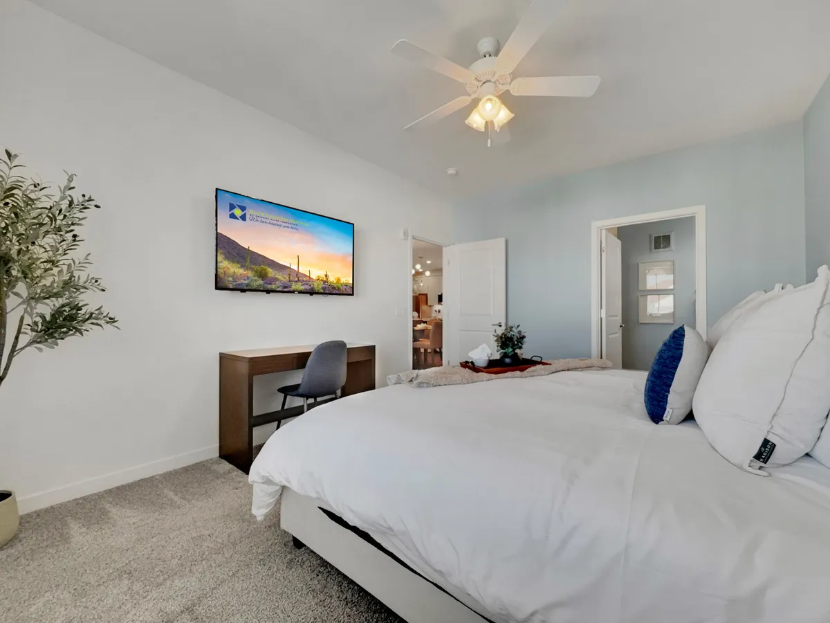 A bright, multi-functional interior room featuring a bed with crisp white bedding, a desk and chair set up for work, a television mounted on the wall, and a touch of greenery. The space is cozy and comfortable, balancing productivity and relaxation.