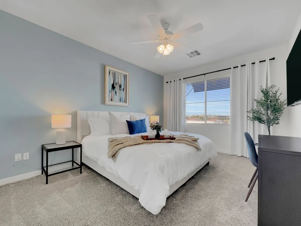 A spacious bedroom with a king-sized bed dressed in white and blue linens, a nightstand with a lamp, a window filling the room with natural light, abstract wall art above the bed, and a desk with a chair set up for work or study.