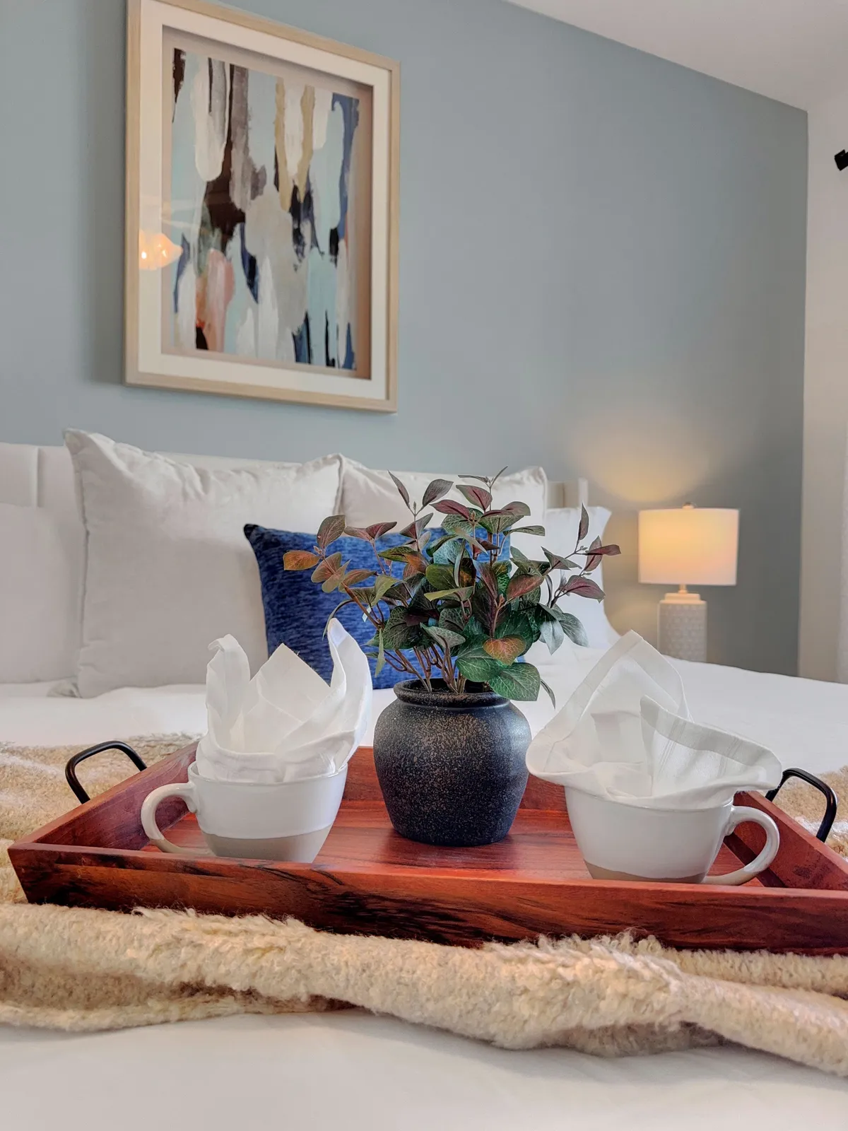 A serene bedroom scene with a wooden tray holding two white cups and napkins atop a neatly made bed, accented by a potted plant and framed artwork. This inviting setup suggests relaxing mornings or unwinding together after a fun day.
