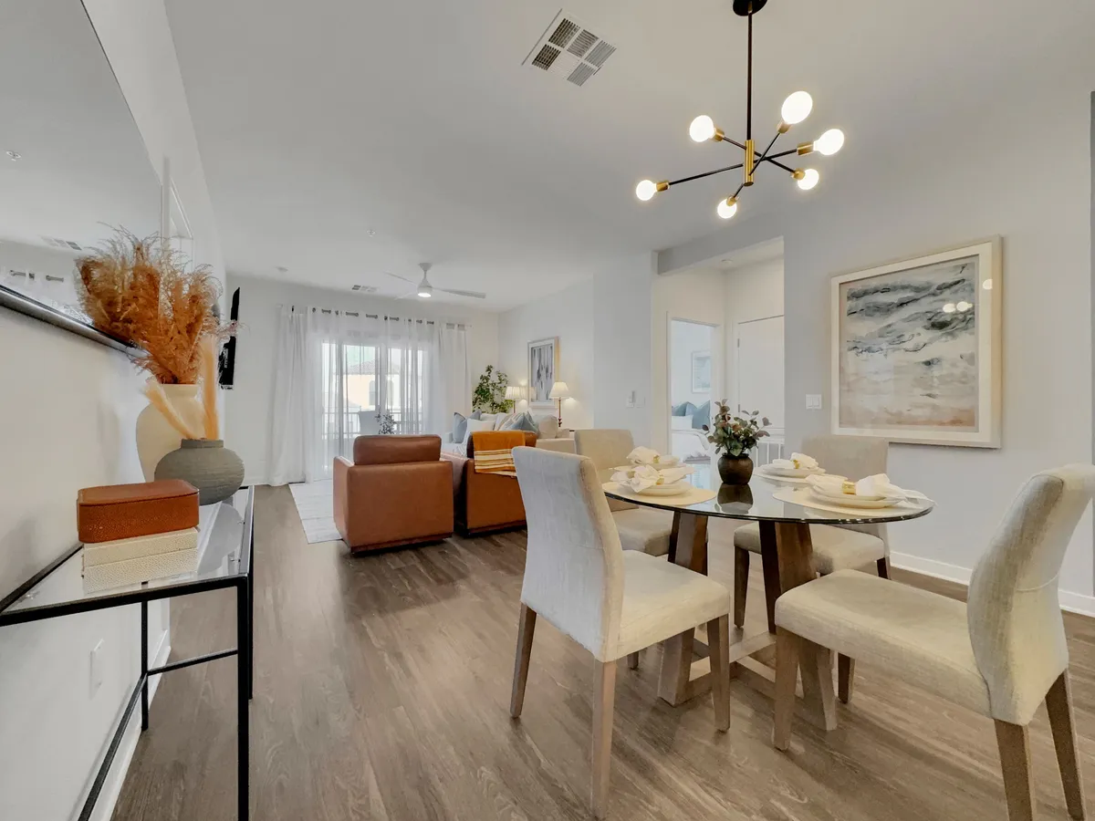A welcoming and contemporary dining area featuring a dining table with comfortable chairs, a statement light fixture above, decorative plants, abstract wall art, and a view of an adjacent cozy seating area. The space is clearly designed for both intimate dining and relaxed socializing.
