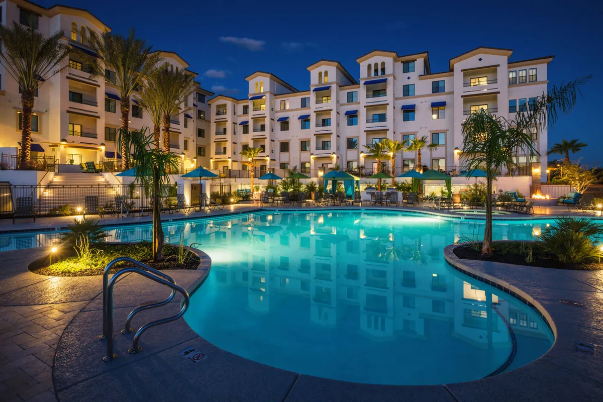 A serene evening view of the community swimming pool, framed by palm trees and illuminated building exteriors. Outdoor seating and gathering areas are highlighted, suggesting both daytime and nighttime use for relaxation and socializing.