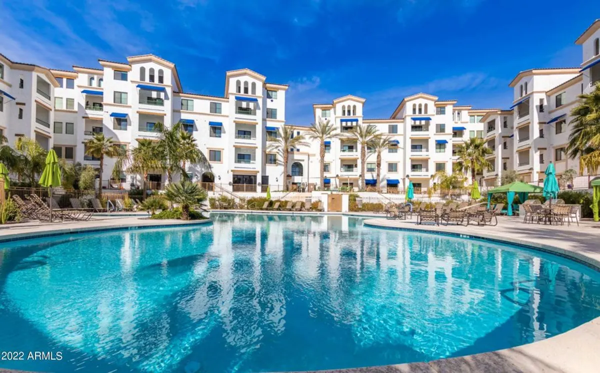 A vibrant exterior scene featuring a large, heated community swimming pool surrounded by tropical palm trees and the condo building facade. Lounge chairs and umbrellas line the deck, suggesting opportunities for sunbathing and socializing. The setting evokes resort-style relaxation and active social gatherings.